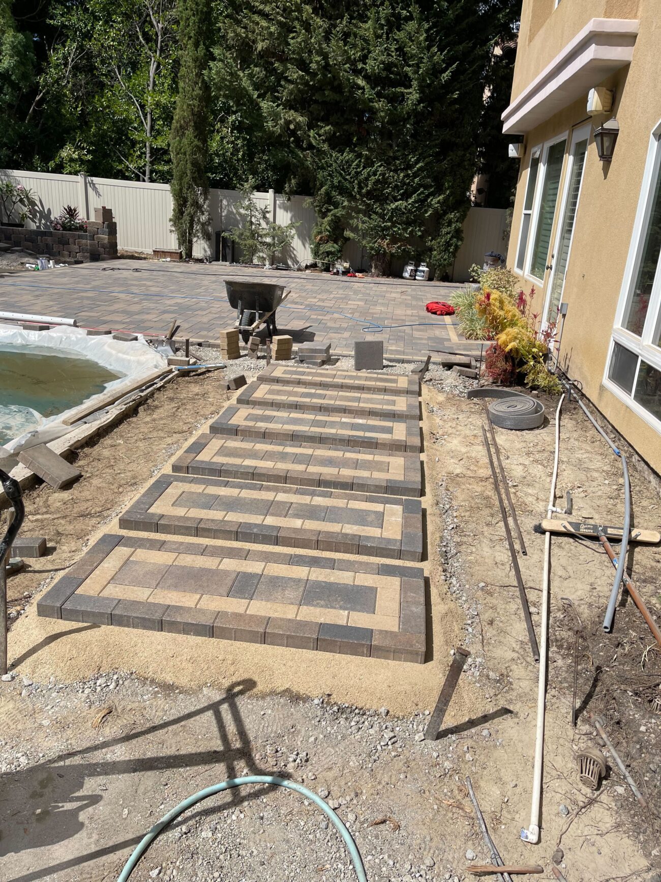 Work-in-progress shot of a residential backyard showing a newly installed interlocking paver walkway leading to a larger...