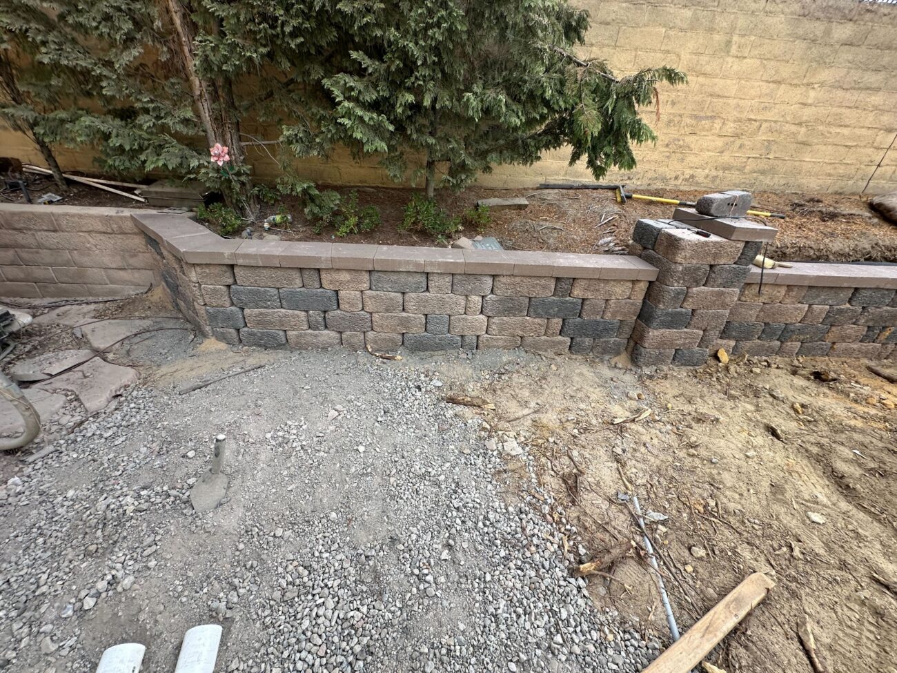 Segmental concrete retaining wall under construction in a residential backyard with capstones in place. A compacted...