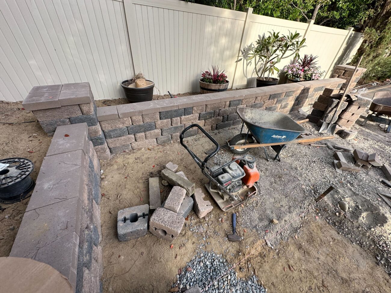 Partial segmental retaining wall built from interlocking concrete blocks runs along a vinyl fence in a residential...