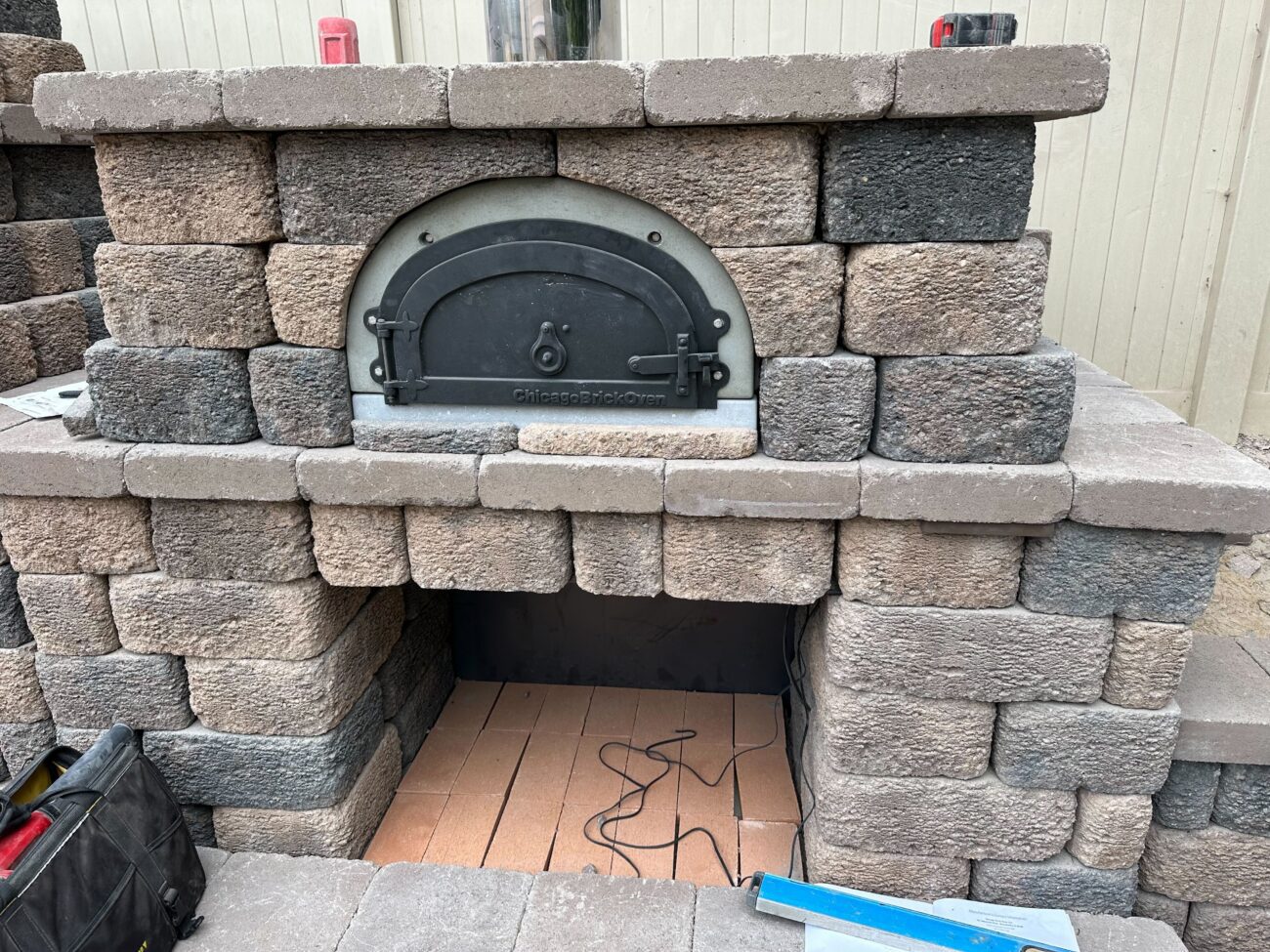 Segmental concrete block outdoor oven structure with a metal oven door installed in the opening. The interior shows a...