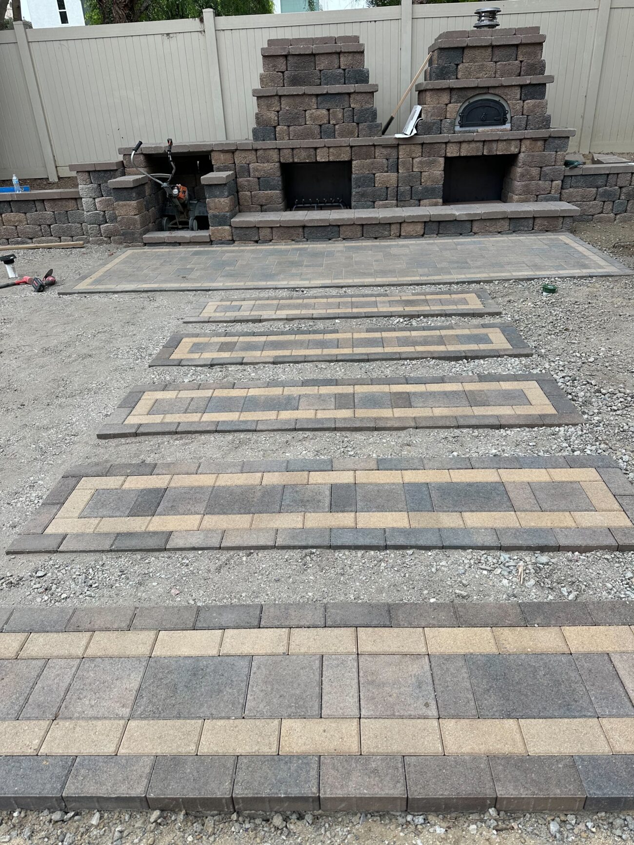 Staged interlocking paver panels laid out on compacted aggregate leading to a partially completed segmental block wall...