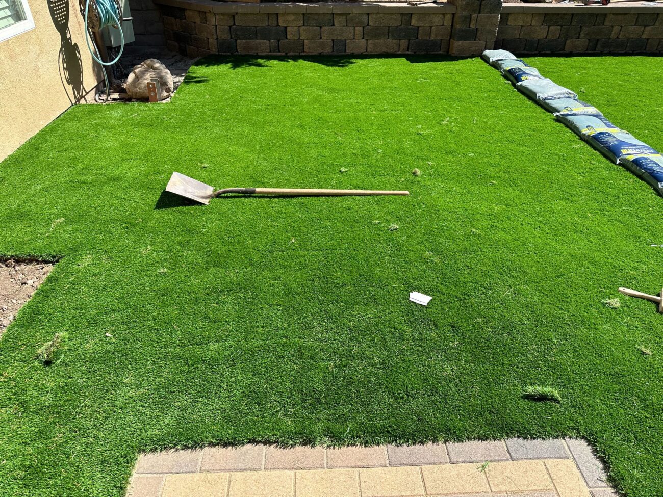 Residential backyard showing an artificial turf installation in progress. Turf rolls are staged along the rear wall and a...
