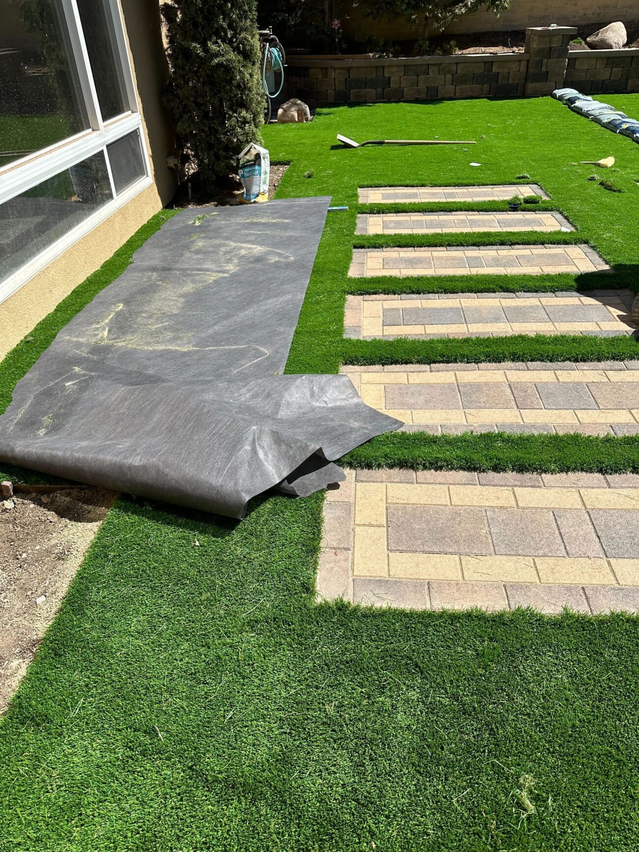 Backyard hardscape installation in progress showing concrete interlocking pavers set as stepping stones with artificial...