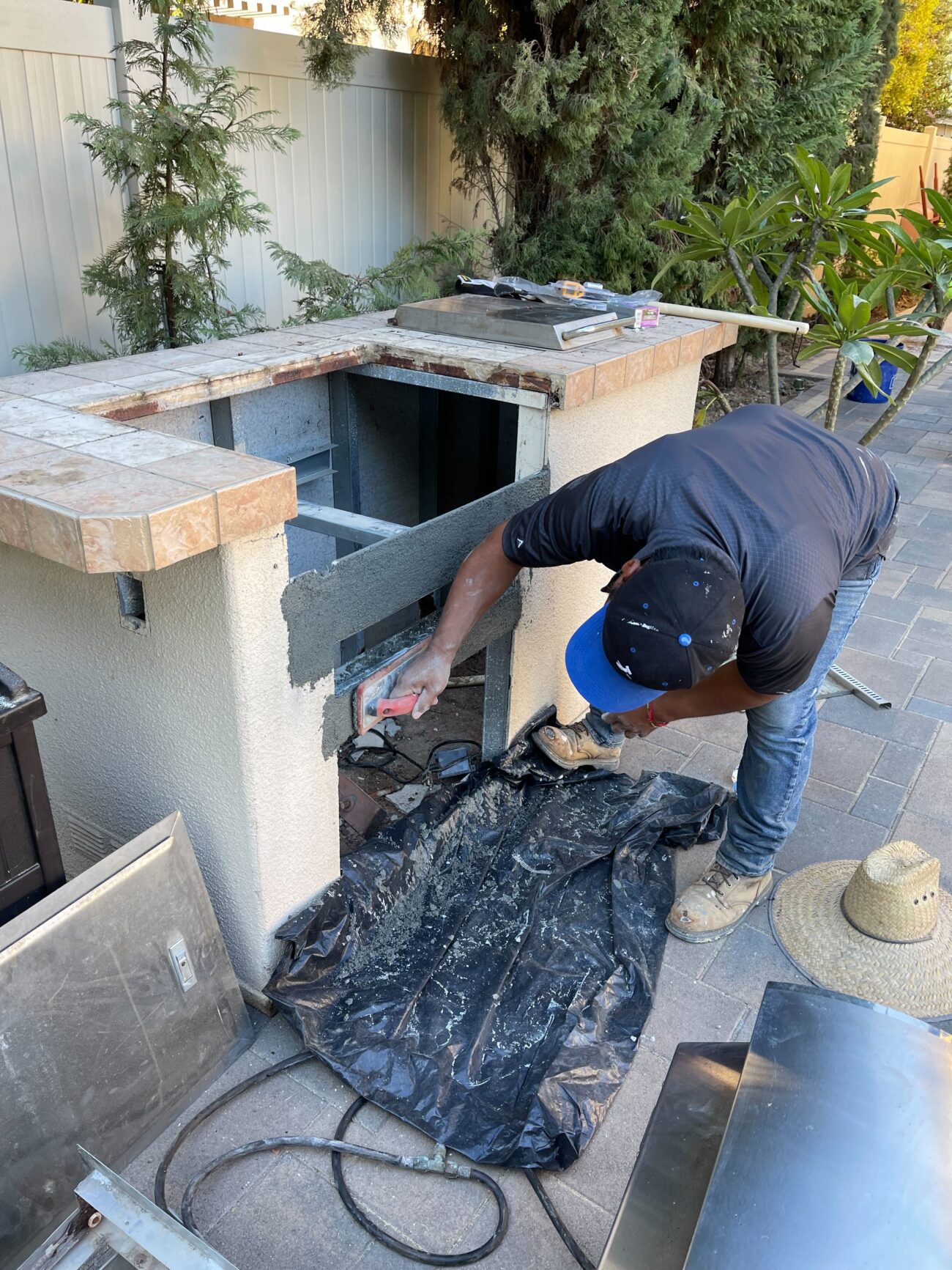 A worker is applying cement/stucco render to the exterior face of a built-in outdoor kitchen island adjacent to an...