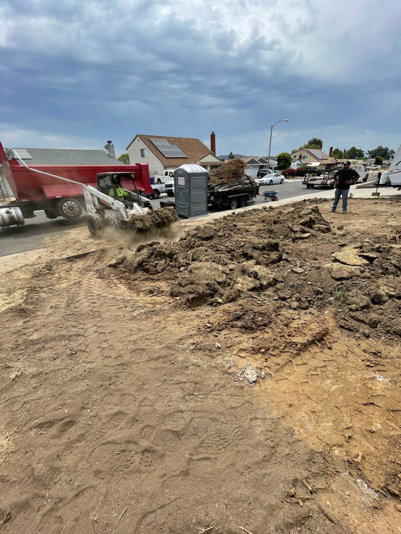 Residential front yard demolition in progress: a skid steer is loading sod and topsoil into a dump truck while crew...