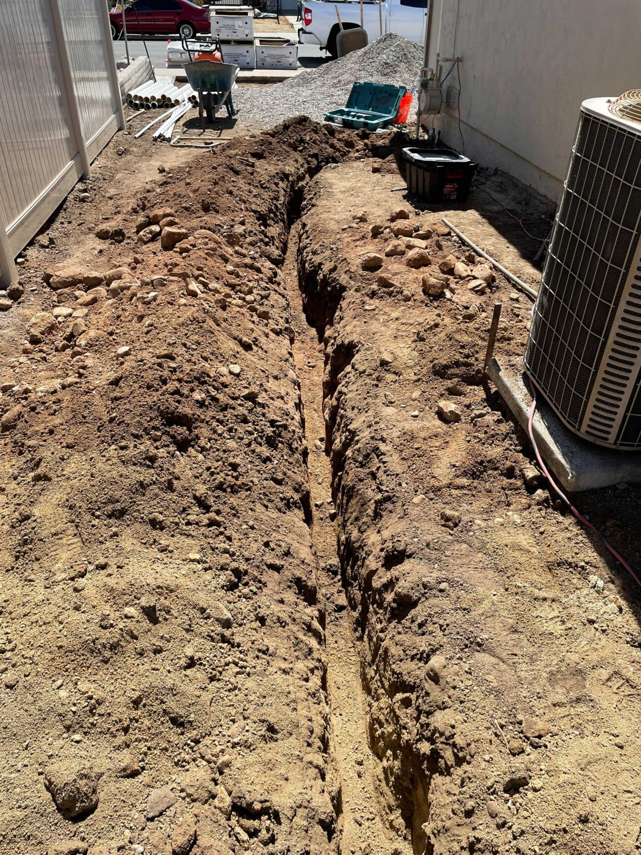 Narrow trench excavated along the side yard between the house and fence, with spoil piles on either side. Appears to be...