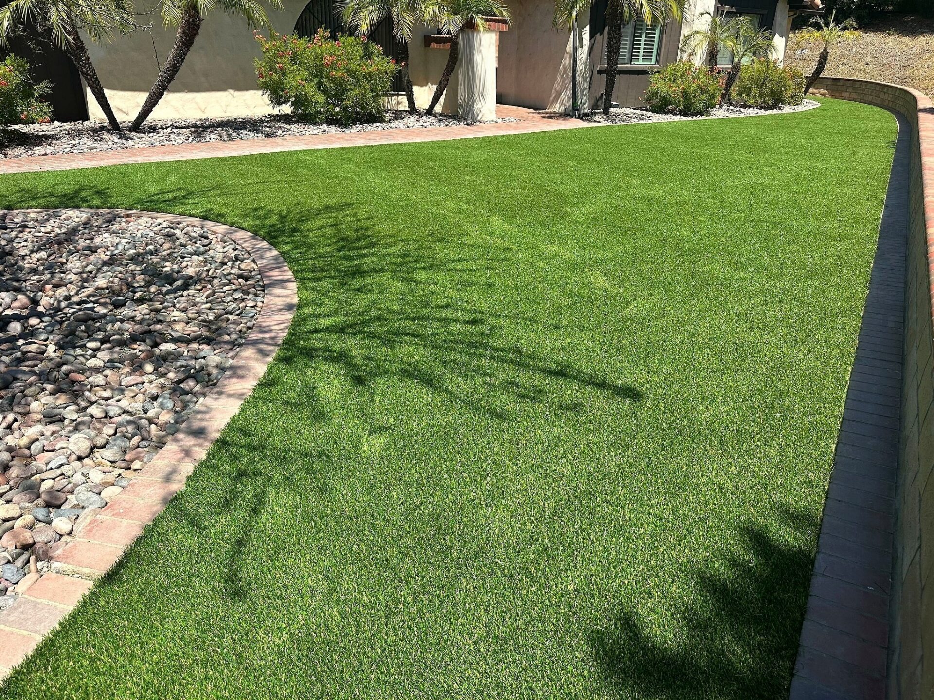 Front-yard SiteOne Rye turf lawn spanning the house facade, framed by a brick-edged river-rock bed and a paver-capped retaining wall.