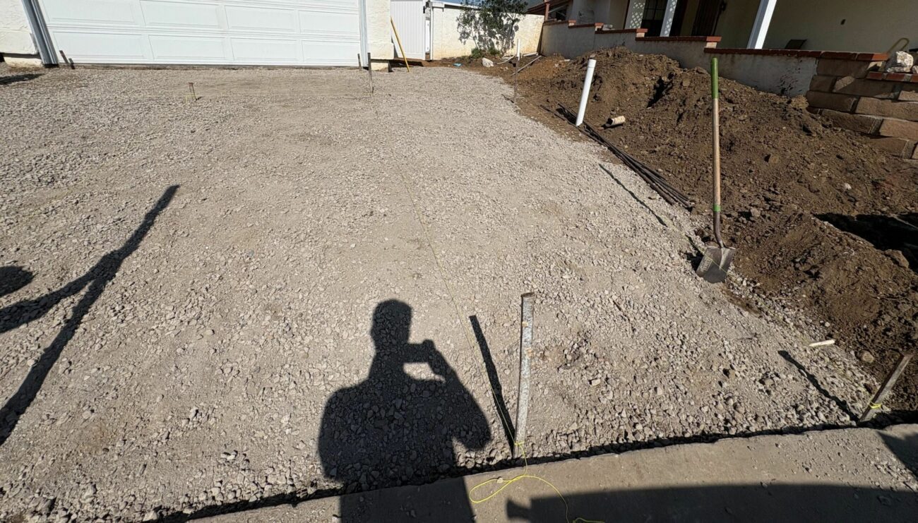 Front driveway graded and compacted with crushed aggregate base, layout stakes marking the paver lines, block retaining wall alongside.