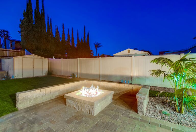 A square gas fire pit with tall flames over lava rock at dusk, surrounded by an L-shaped block seat wall on an interlocking-paver patio, with a turf lawn and cypress trees behind.