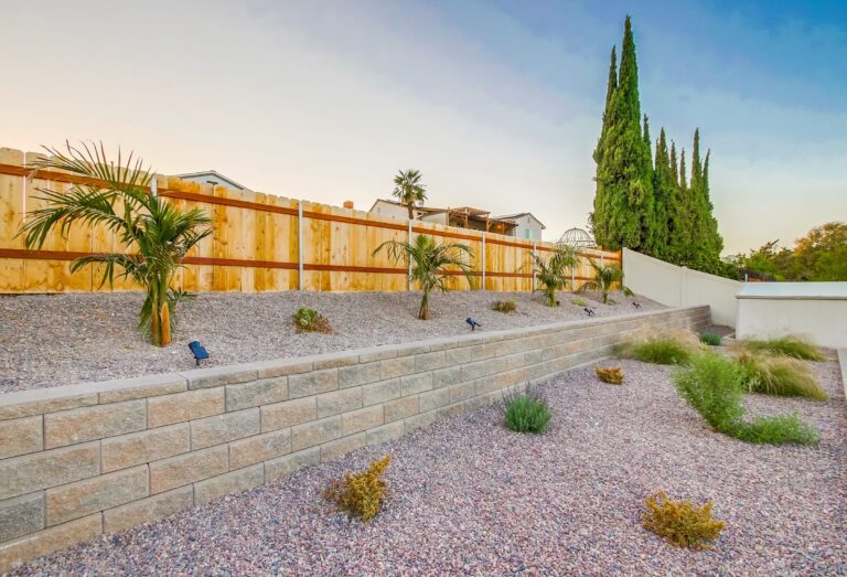Angled dusk view of the multi-tier retaining-wall garden, gravel planting terraces with grasses and shrubs stepping up to a wood fence, cypress trees at right.