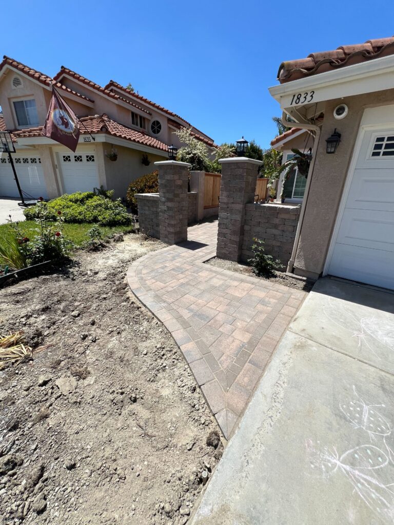 Front entry approach from the driveway: a curved mixed-Angelus paver walkway with a soldier-course border rising between two stone-veneer pillars to the courtyard gate of a Vista home.