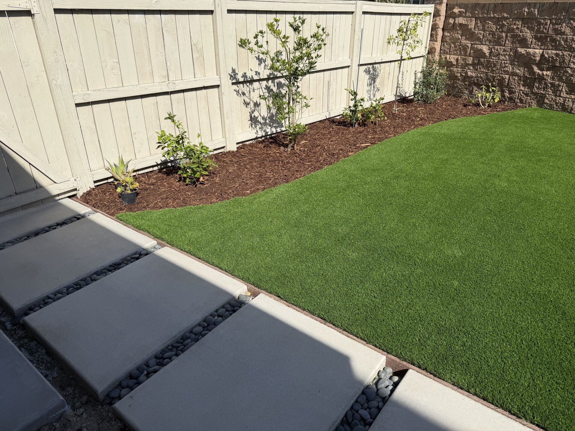 Synthetic turf beside a curved mulch bed with new plants and concrete step pads set in black beach pebble, Escondido 92029