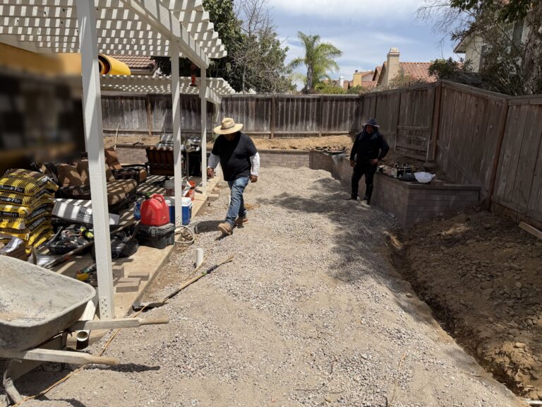 Build stage: crew spreading compacted base for the turf after the wall went up, with the drain line trench open along the fence